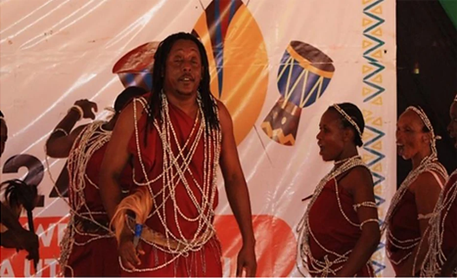 A Taveta performer in red attire and long bead necklaces dancing the Uruasi on stage.